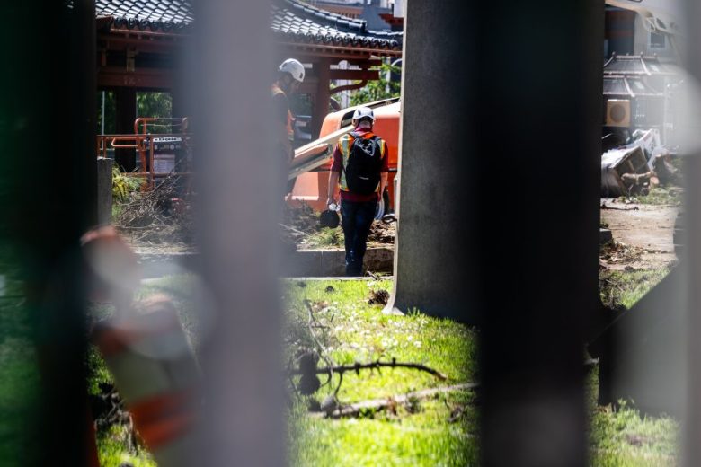 Two construction workers in safety gear stand near equipment at an outdoor worksite, seen through vertical metal bars with scattered debris on the ground.