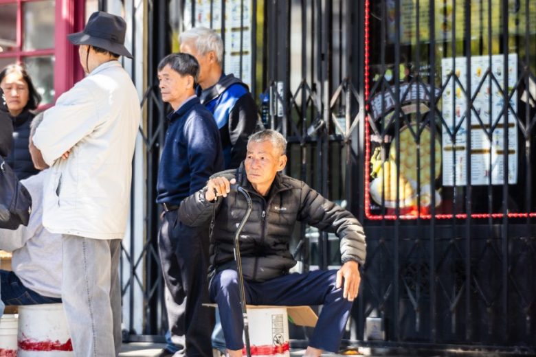 Several people stand and sit outside a storefront; an older man with a cane sits in the foreground, while others stand and talk nearby.