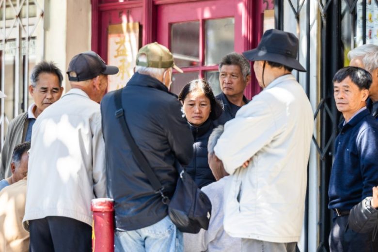 A group of people stands and sits closely together outside a building with red doors, engaged in conversation on a sunny day.