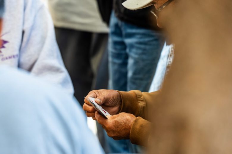A person wearing a cap and brown jacket holds a hand of playing cards while sitting among a group of people.