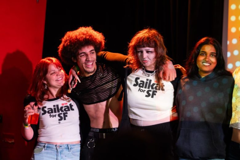 Four people stand closely together smiling; two wear matching "Saikat for SF" shirts. The background is a dark stage with red and yellow lighting.