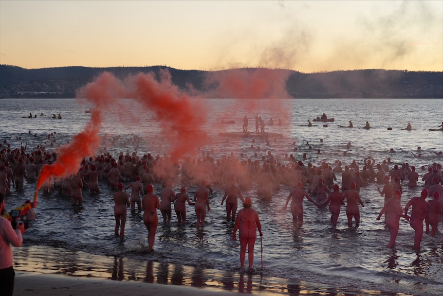 a crowd of naked people run into the river as red flares are shot into the air.