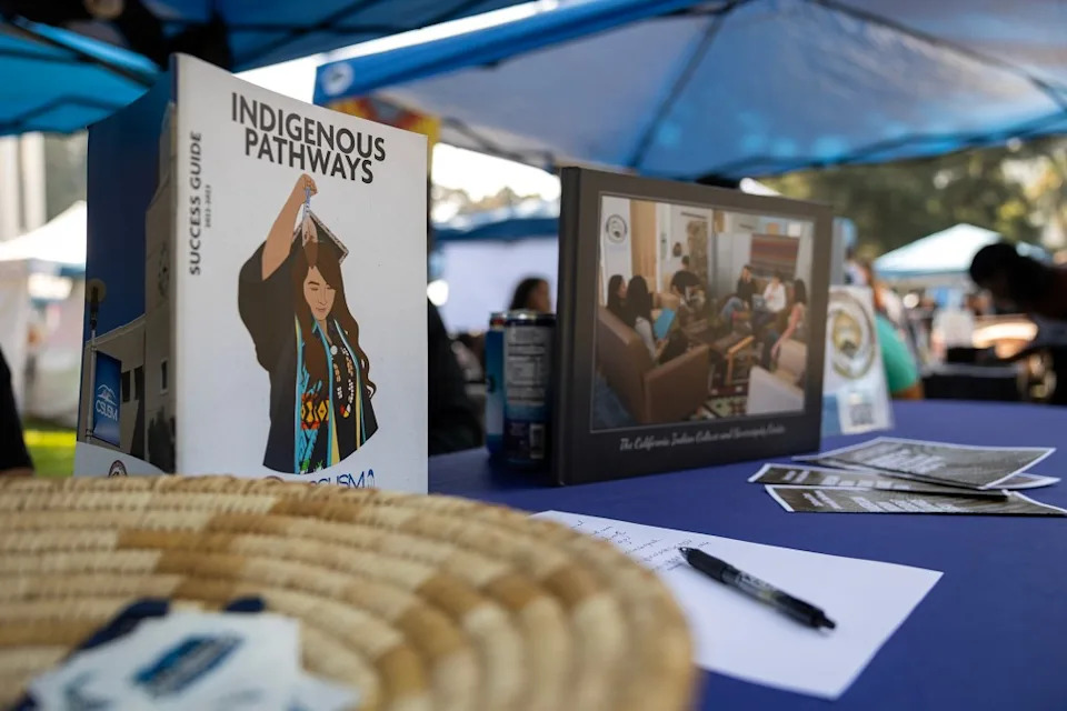 A table display under blue canopy tents features a standing booklet titled “Indigenous Pathways” with an illustrated graduate on the cover. Nearby are a framed photo, printed flyers, a pen resting on a sign-in sheet, and a woven basket blurred in the foreground, while people and booths appear softly out of focus in the background.