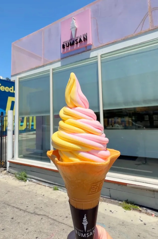 Pink and yellow soft serve ice cream in a cone, with a pink building that reads “Organic Milk Bar Bumsan” in the background Instagram@bumsanorganicmilkbar