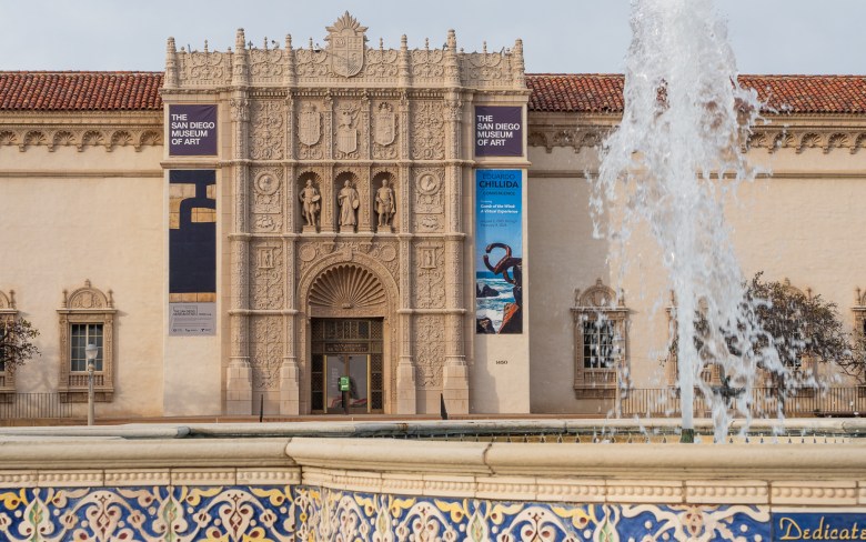 The Plaza de Panama fountain spouts water in front of The San Diego Museum of Art. Decorative tiles line the walls of the fountain. Three statues, portraits, shields, and other icons are displayed in the ornate facade of the Museum of Art.