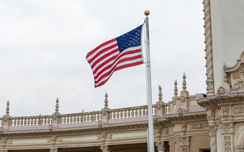 An American flag billows in the wind on a bright, cloudy day. The ornate walls and railings of the Spreckles Organ Pavilion line the bottom and right sides of the image.