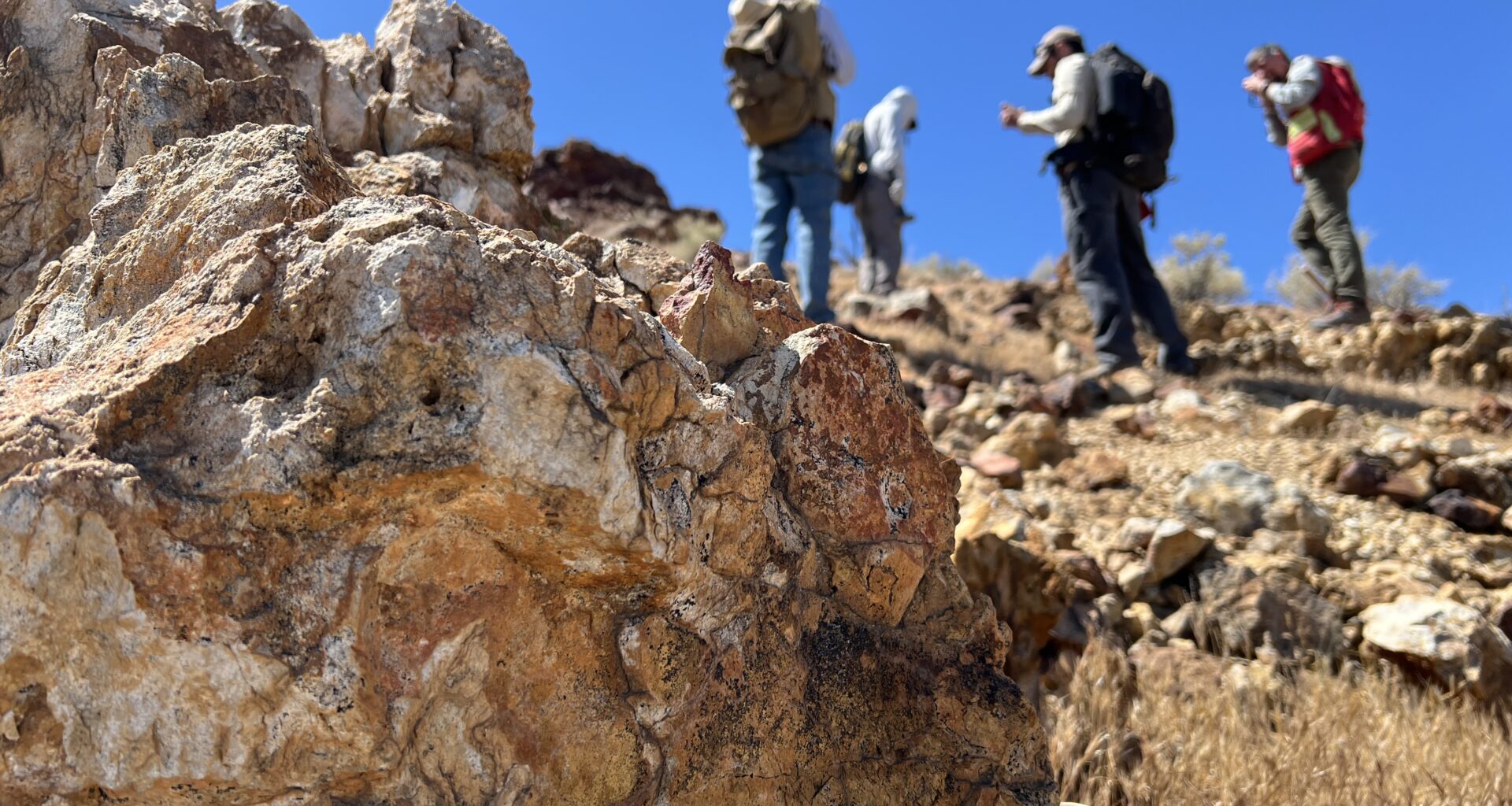 Four people with backpacks pause to examine rocks on a hillside under a clear blue sky, with a large orange-and-tan boulder in the foreground.