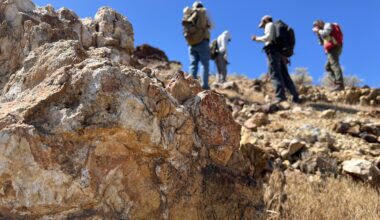 Four people with backpacks pause to examine rocks on a hillside under a clear blue sky, with a large orange-and-tan boulder in the foreground.