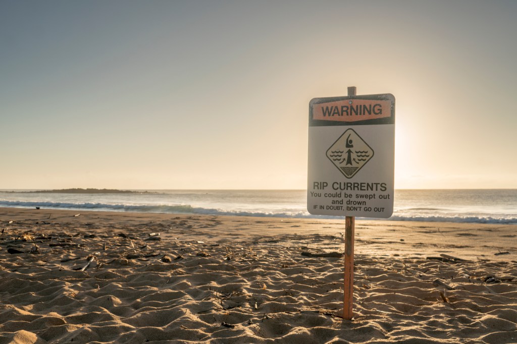 A rip currents warning sign on a sandy beach in Hawaii.