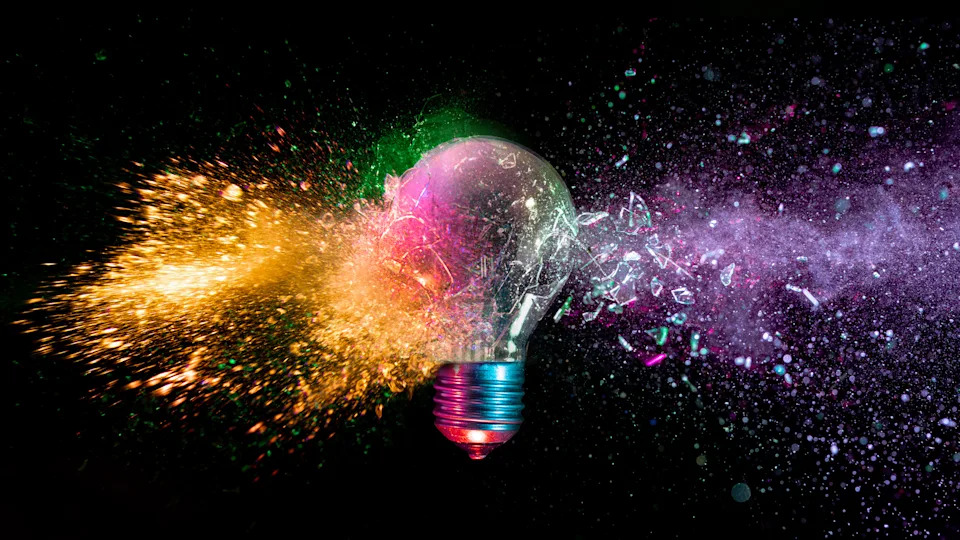 A lightbulb is shattered with various colors coming out of it against a black background