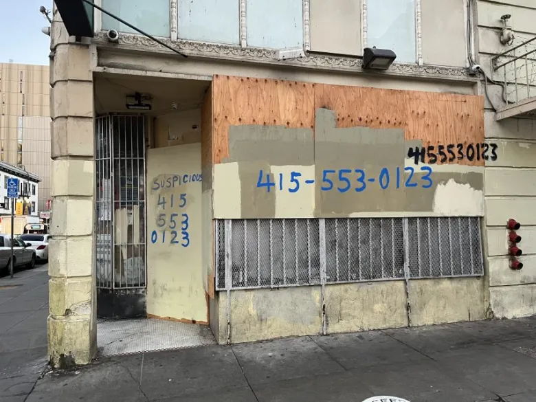 A boarded-up building with metal bars on the entrance and "415-553-0123" painted multiple times on plywood and walls, along with the word "SUSPICIOUS.