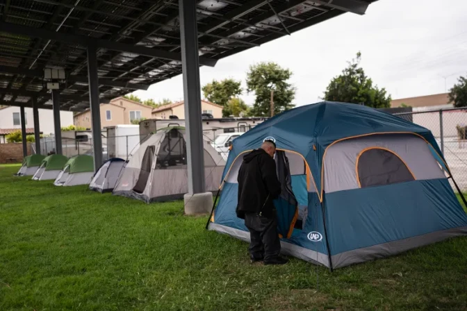 A man stands outside a blue tent next to other tents under a metal structure.