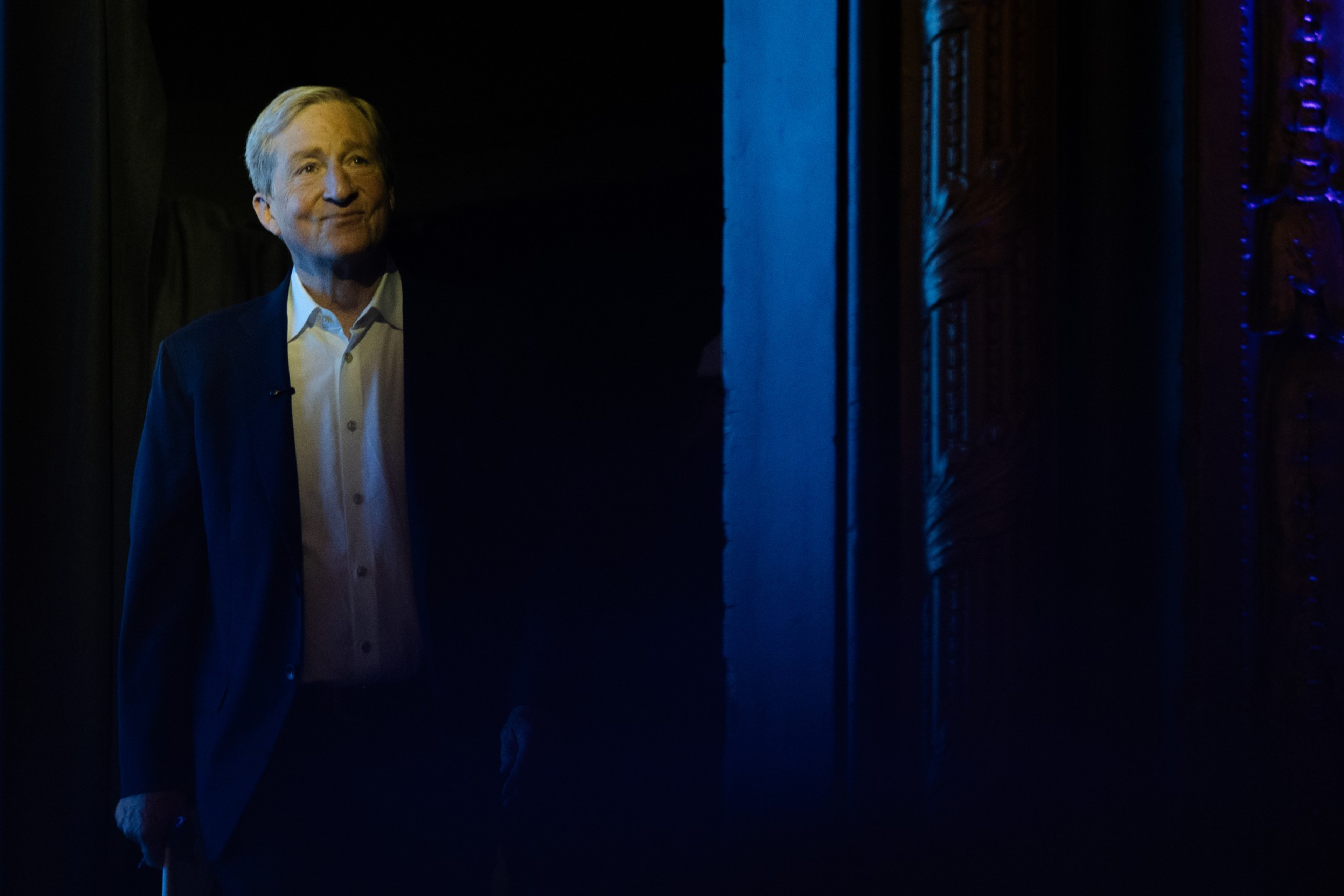 An older man in a dark suit and white shirt stands partially lit in blue light, with a faint smile, against a mostly dark background.
