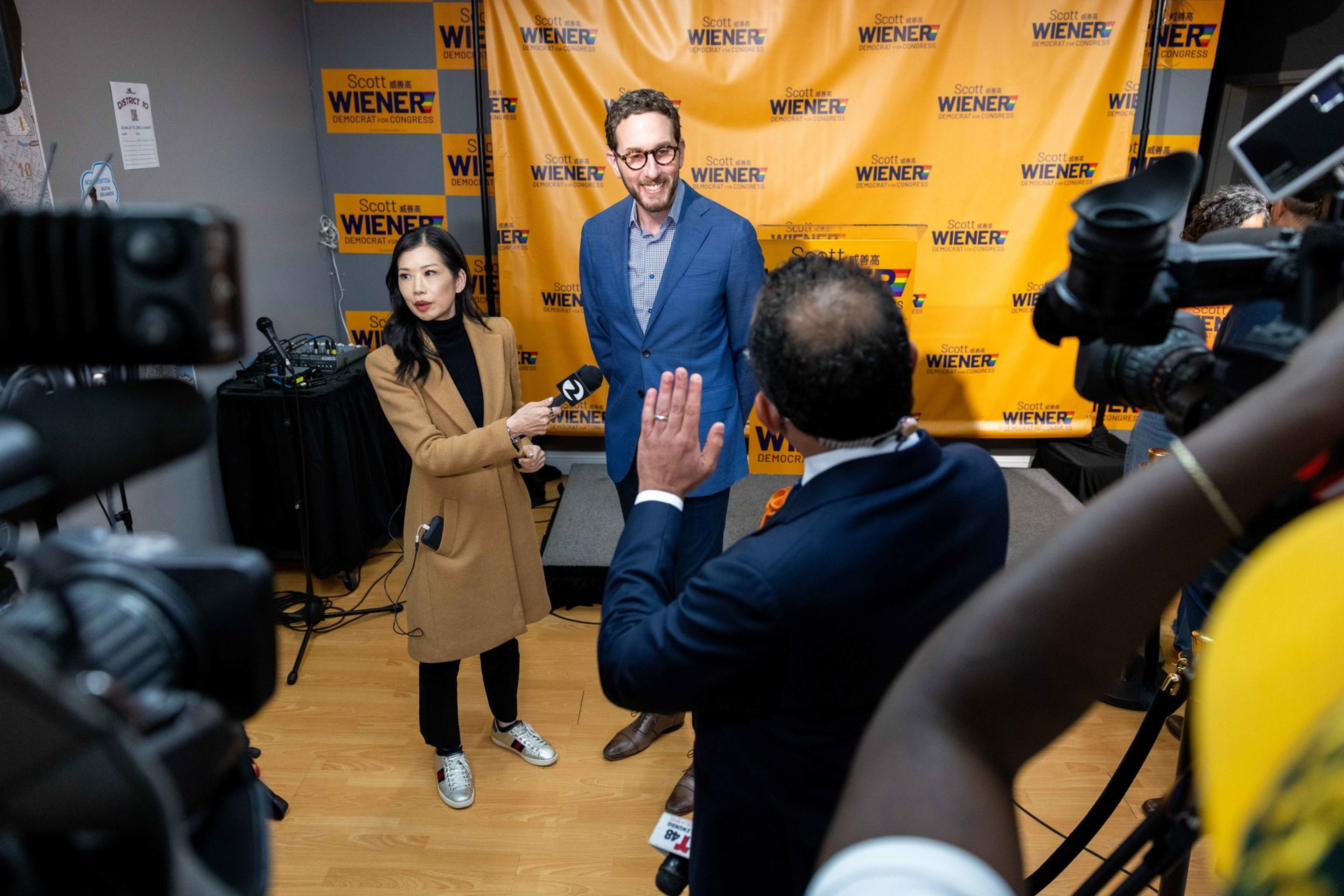 A man in a blue suit stands smiling before a yellow backdrop with “Scott Wiener” logos while a female reporter holds a microphone toward him amid camera crews.