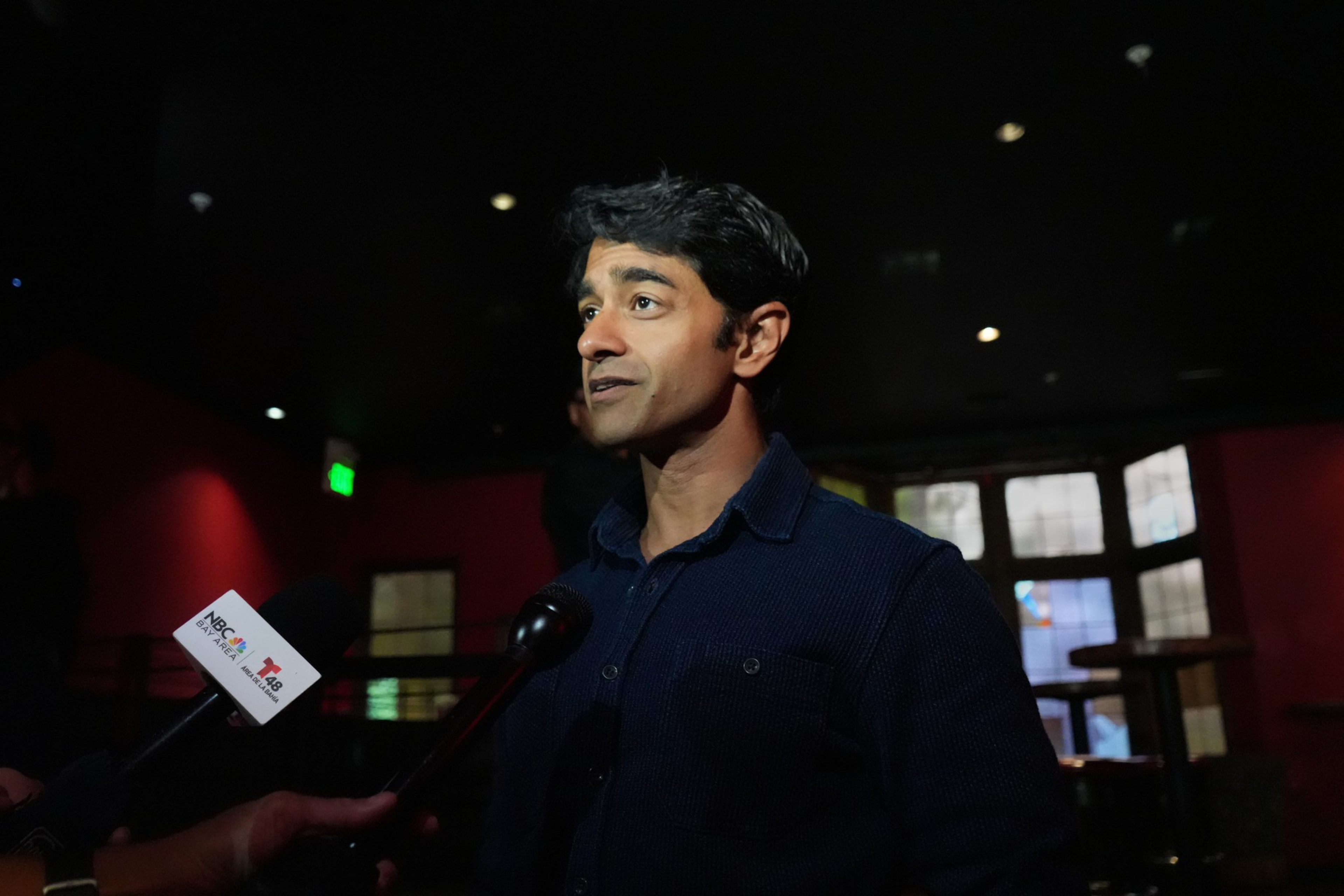 A man with dark hair wearing a dark shirt is speaking to reporters holding microphones in a dimly lit room with windows behind him.