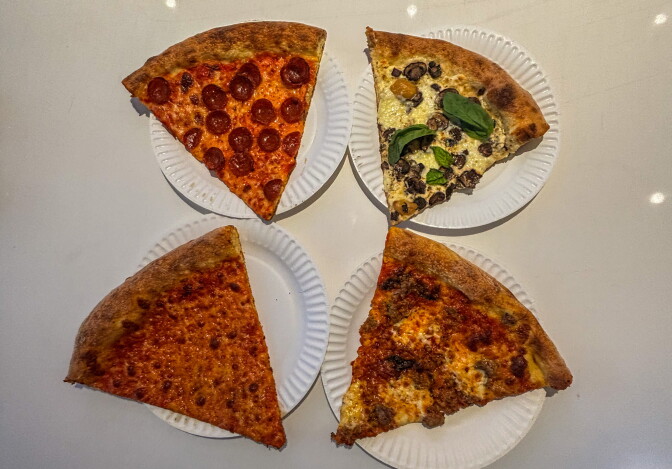 Four pizza slices on white paper plates arranged in a square on a white surface: clockwise from top left, a pepperoni slice, a white mushroom slice with fresh basil, a sausage slice, and a plain cheese slice.