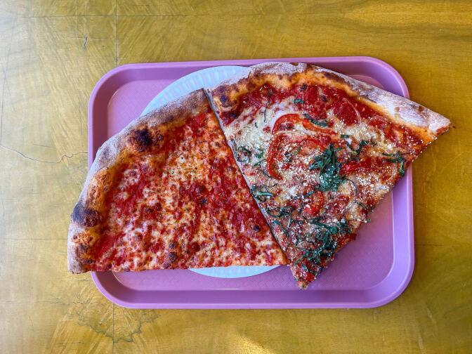 A pink plastic tray sits against a light brown background. On the tray is a partially shown white paper plate. On the left is a large cheese slice dotted with red sauce; next to another large slice contains red vegetables and cut basil baked into the red sauce and white cheese.