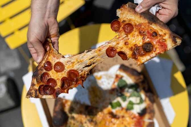 A round pizza with four different kinds of slices: one is cheesy with round pepperoni, next to it a slice with chunks of white burrata and green basil leaves, then a pizza with red sauce, and another cheese slice with a green pesto-looking sauce. The pizza is on a top of an open pizza box which sits on a bright round yellow table atop asphalt of a parking lot. Two hands hold the pepperoni slice and split it up so the melted cheese is the only thing adjoining them.