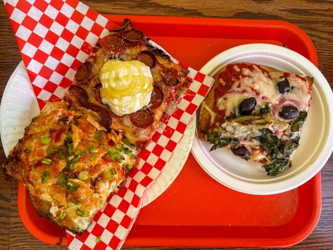 Three pizza slices on a red cafeteria tray: a square slice topped with pepperoni and a dollop of burrata on red checkered paper, a triangular slice with crab and scallions on a paper plate, and a round slice with olives, ham, and greens on a white plate.