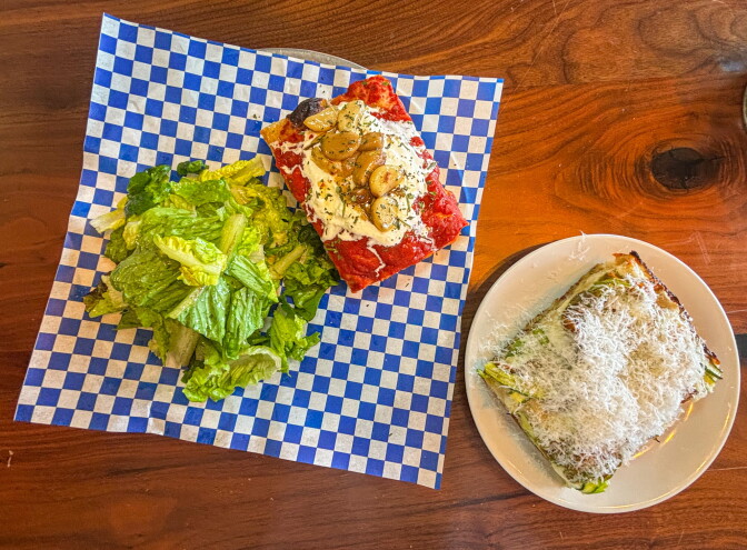 Two square focaccia slices and a side house salad on blue checkered paper and a white plate on a wood surface. One slice is topped with confit garlic, ricotta, and tomato sauce; the other is a cacio e pepe style slice blanketed in finely grated cheese.