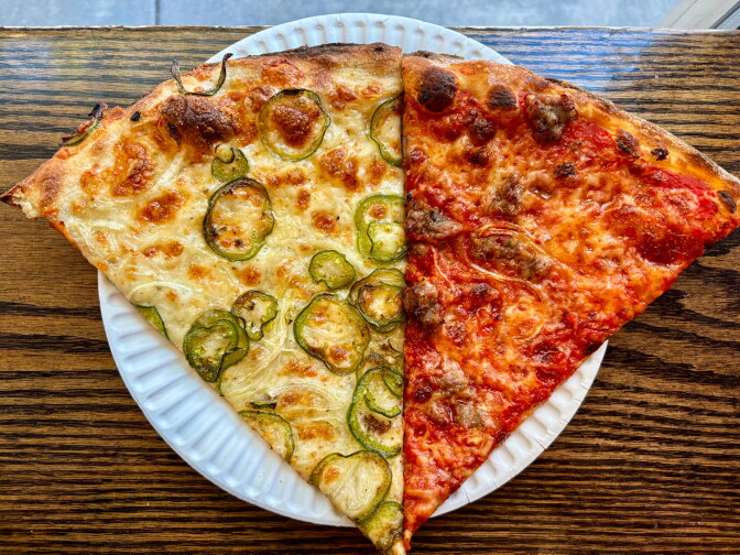 Two large pizza slices side by side on a white paper plate — one topped with jalapeño rounds and melted cheese, the other with tomato sauce, melted cheese, and bits of sausage — sitting on a wooden table.