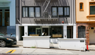 A gray building with black-framed windows and a fire escape has a white low wall with "1030" on it, next to a yellow building and a parked black car.