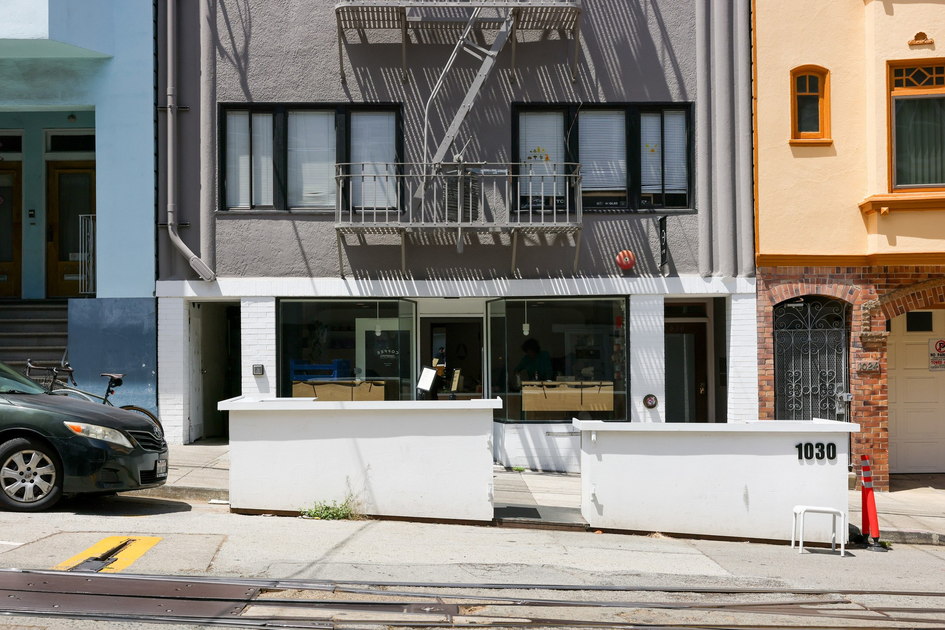 A gray building with black-framed windows and a fire escape has a white low wall with "1030" on it, next to a yellow building and a parked black car.