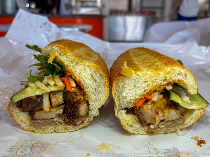 A heo quay banh mi cut in half and laid open, revealing chunks of crispy pork belly, shredded daikon and carrots, cucumber spears, and fresh cilantro on a soft baguette.