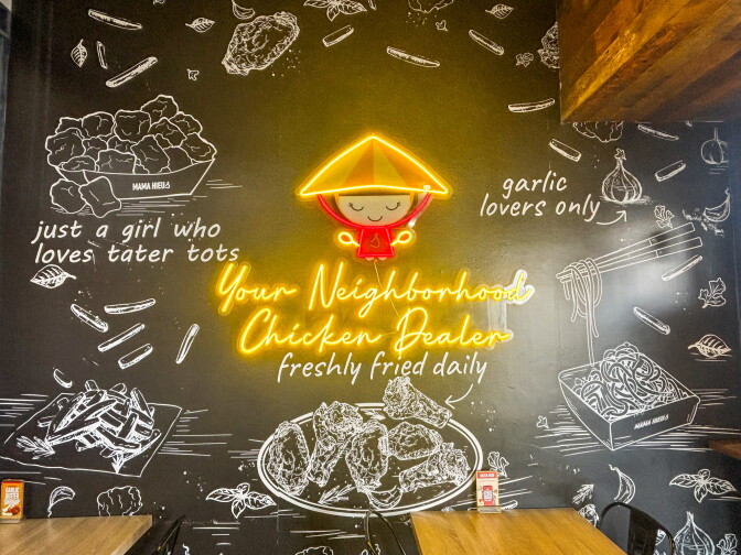 The interior mural at Mama Hieu's featuring a glowing yellow neon sign that reads "Your Neighborhood Chicken Dealer — freshly fried daily," surrounded by chalk illustrations of wings, noodles, and fries on a black wall.