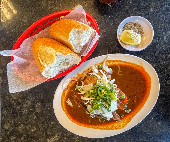 An overhead view of a bowl of bò kho — Vietnamese beef stew — in a deep crimson broth topped with jalapeño rounds, white onion, and green onion, served alongside a basket of sliced baguette and a lemon wedge.