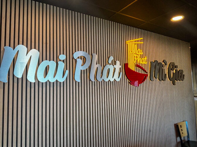 The interior logo wall at Mai Phát Mì Gia, featuring the restaurant's name in flowing white script with a glowing yellow neon noodle bowl emblem against a wood slat backdrop.