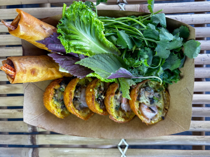 An overhead view of a combo box from Bánh Khọt Lady containing five golden bánh khọt — crispy rice flour pancakes topped with shrimp — two crispy egg rolls, and a bundle of fresh herbs including cilantro, purple Thai basil, and lettuce.