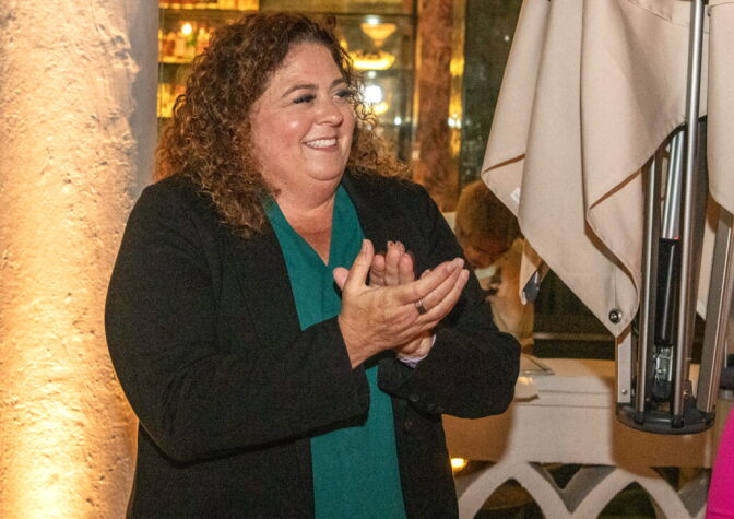 District 5 Councilmember Megan Kerr, a woman with light skin tone, curly hair, wearing a black jacket over a teal shirt, smiles and claps as she looks out of frame.