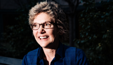 A smiling person with short, wavy hair and glasses is outdoors, wearing a blue shirt and vest, with dark foliage in the background.