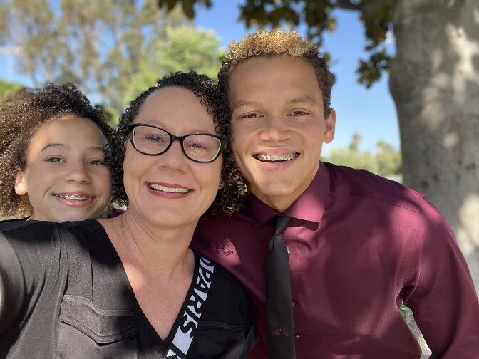 A woman with light skin tone and curly short hair takes a selfie between two children, a girl at left and a boy at right. 