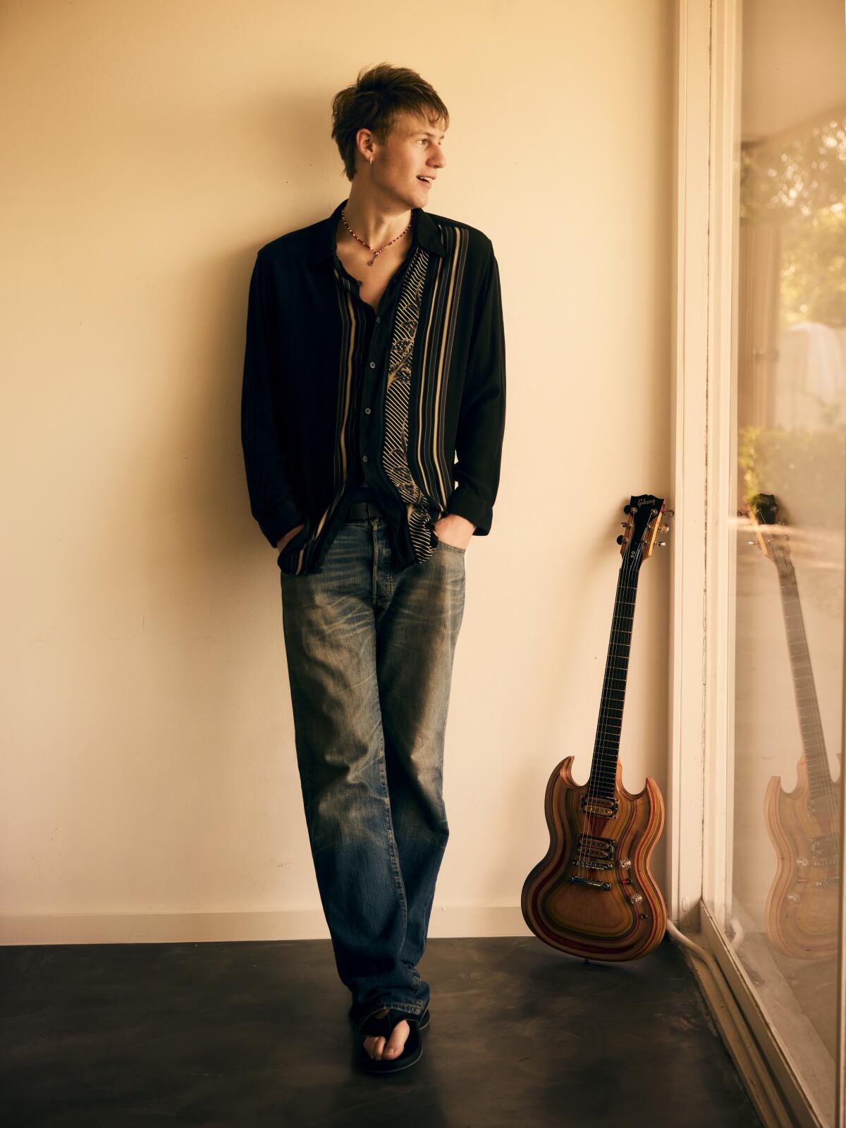 Man leaning against a wall next to a guitar. 