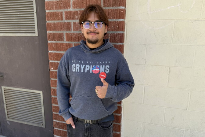 A young man with medium skin tone wears a gray sweatshirt and red, white and blue "I voted" sticker. He gives a thumbs up with his left hand.