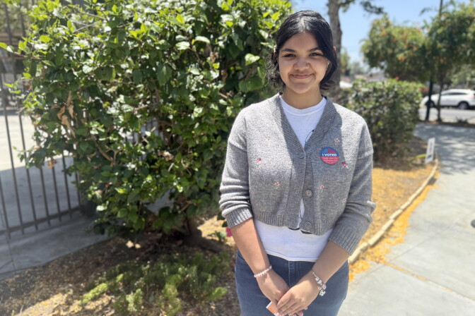 A woman with medium skin tone wears a gray sweater over a white shirt and a red, white and blue "I voted" sticker.