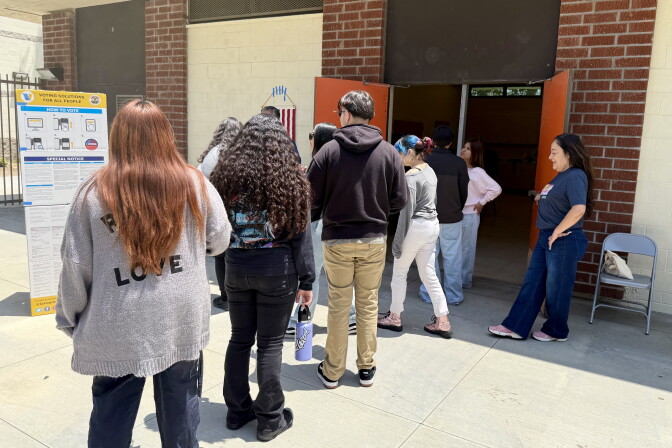A line of young people wait outside the open doors of a brick building.