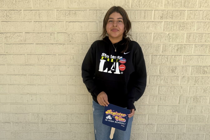 A young woman with medium skin tone wears a black sweatshirt and a red, white and blue "I voted" sticker.