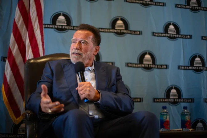 Former Gov. Arnold Schwarzenegger, a man with medium skin tone, brown short hair, and a gray beard, wearing a blue suit, speaks into a microphone as he sits and looks out of frame.