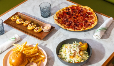 A pepperoni pizza, a burger with fries, a salad with croutons and lemon, and a plate of stuffed potato skins with dipping sauce are set on a table.