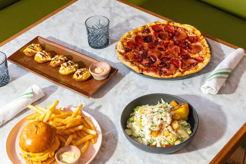 A pepperoni pizza, a burger with fries, a salad with croutons and lemon, and a plate of stuffed potato skins with dipping sauce are set on a table.