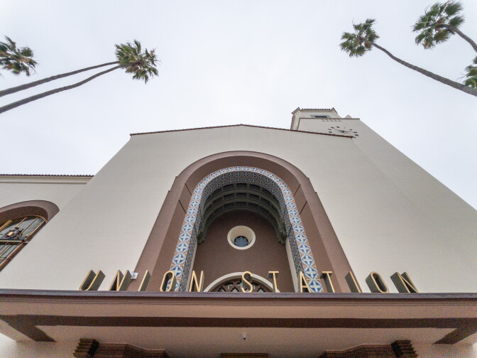 This is a photo of Union Station. A view looking upward of a cream colored building with large brown arch way. Scenery of four palm trees on the side of the building.