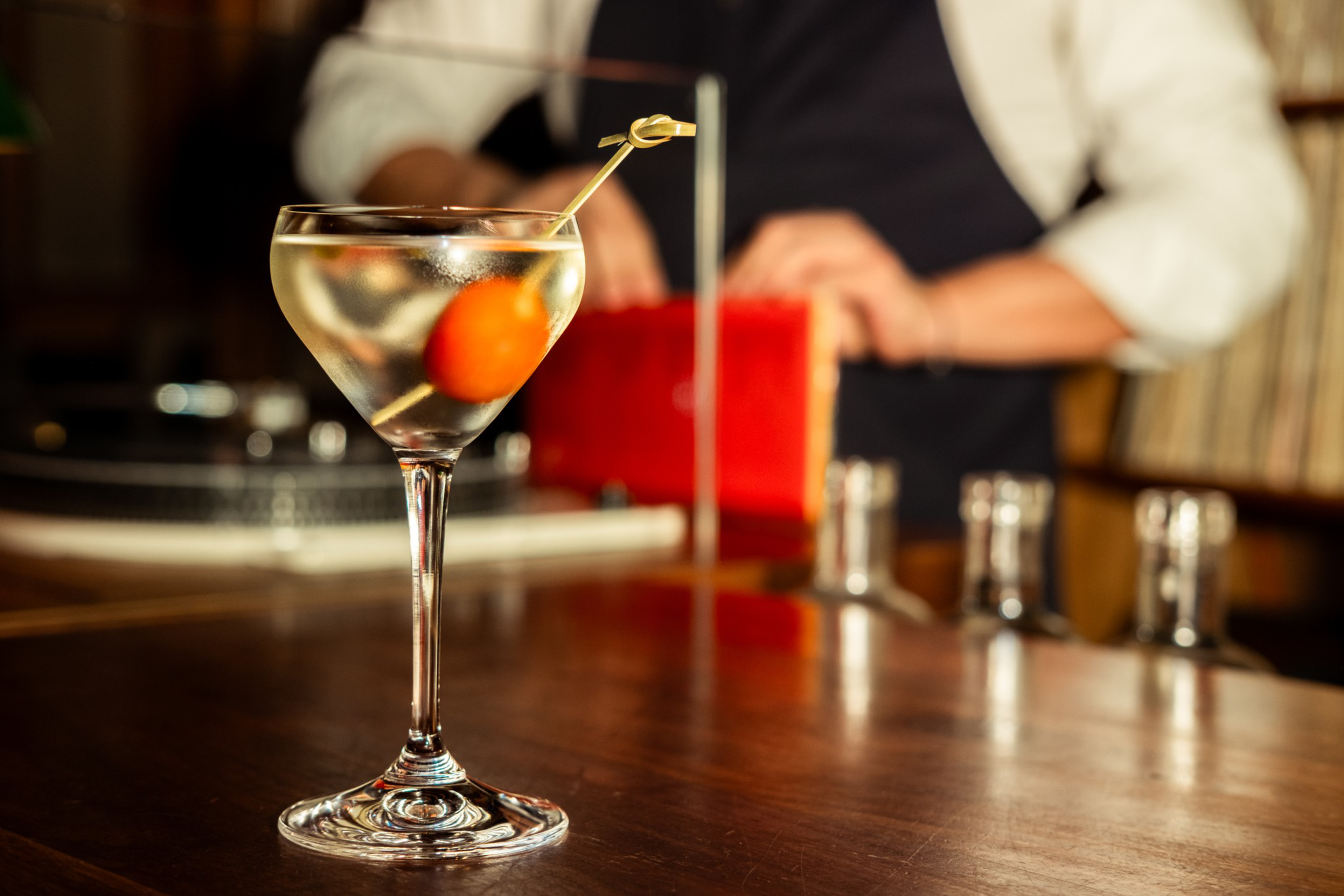 A clear cocktail in a stemmed glass garnished with an orange cherry on a pick sits on a wooden bar counter.