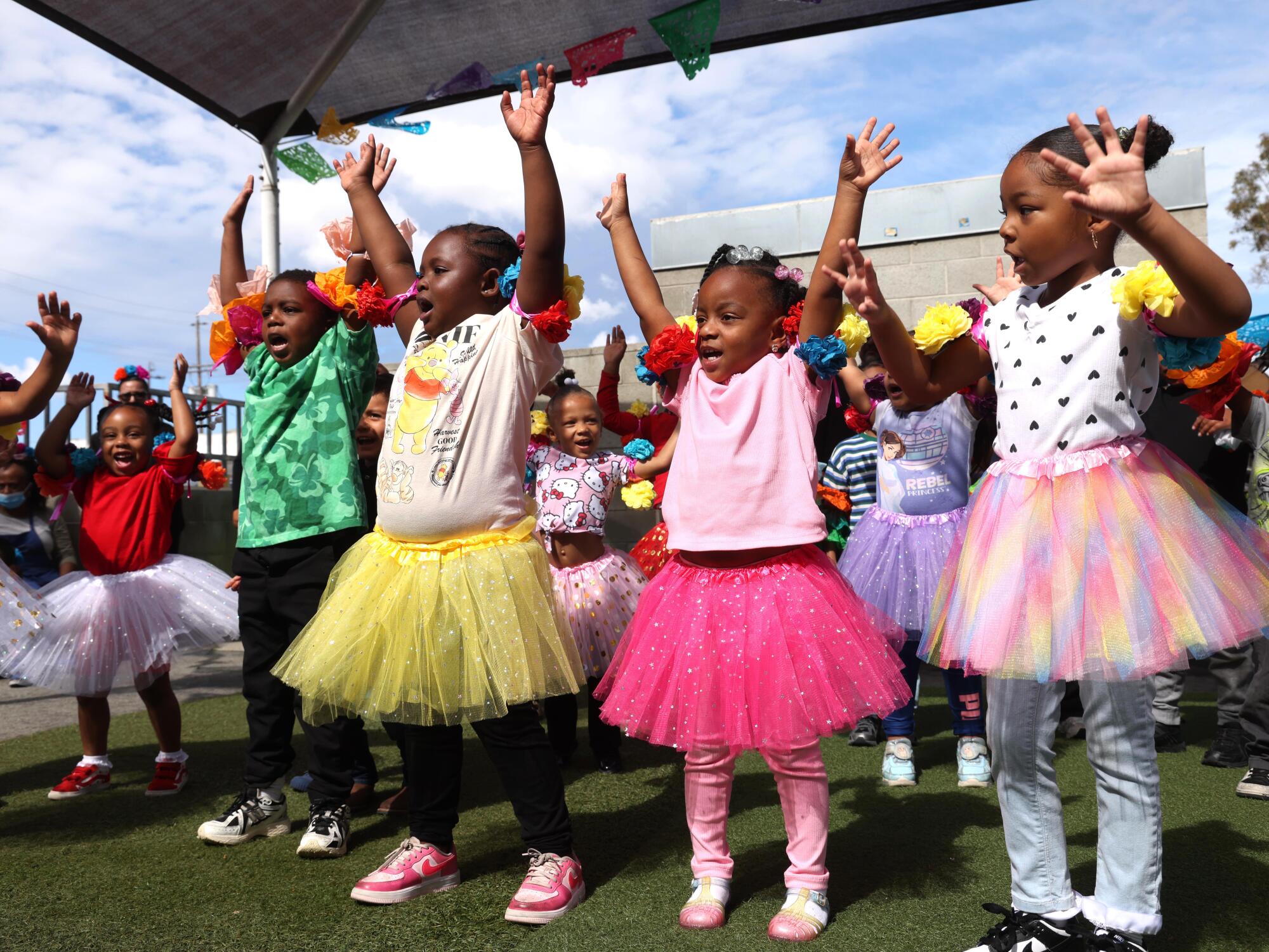 Preschoolers wearing tutus raise their hands in the air during an outdoor dance performance for Cinco de Mayo.