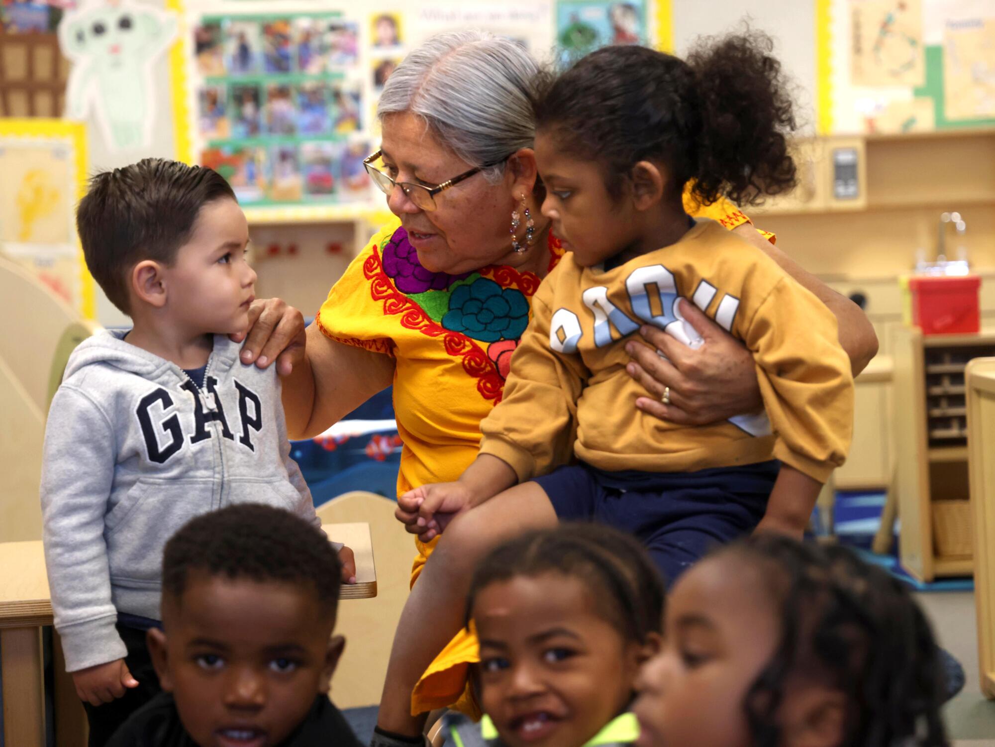 A woman holds one child in her lap and chats with another as students gather on the classroom rug.