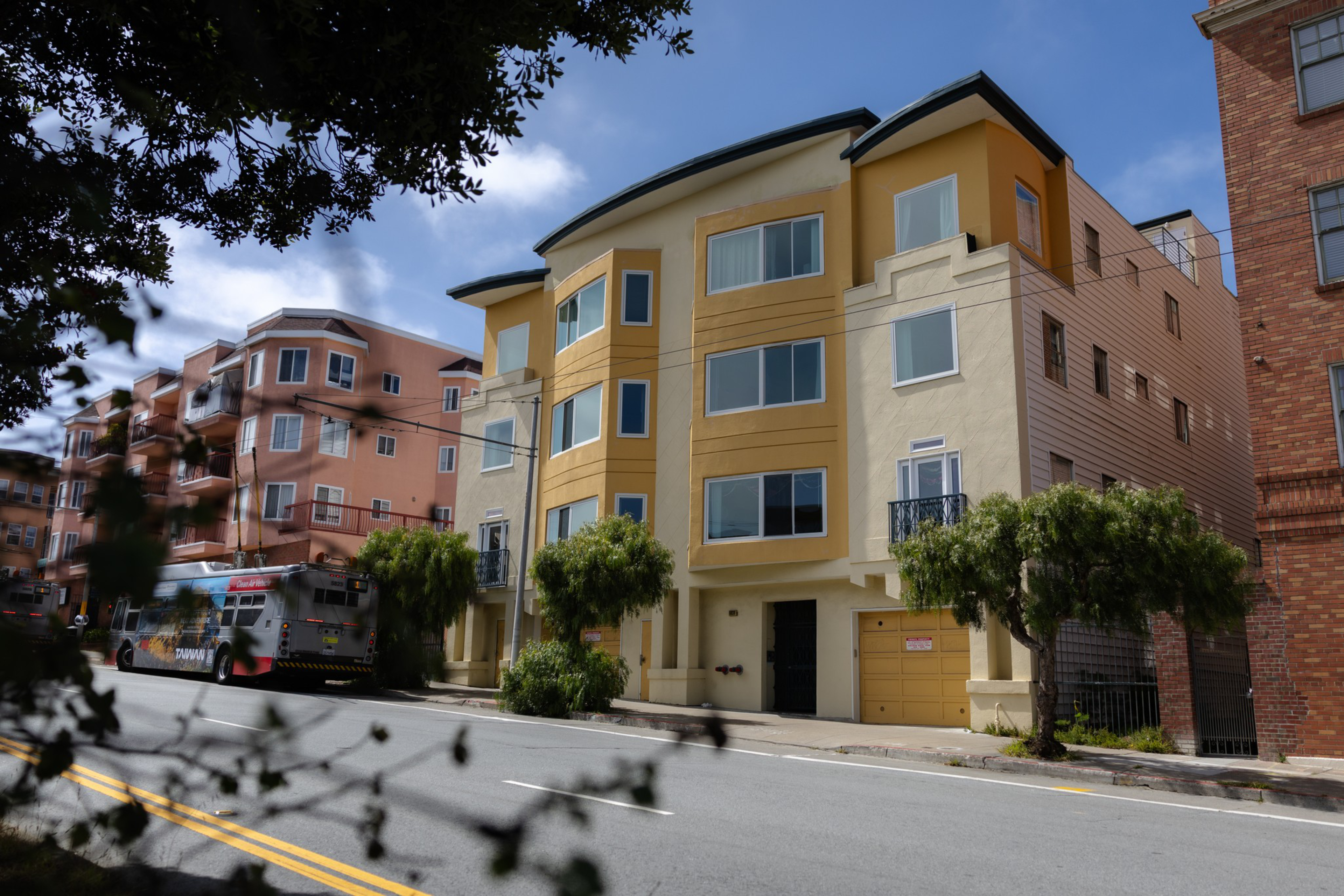 A yellow and beige three-story apartment building sits on a street with small trees in front, next to a bus and another multi-story building.