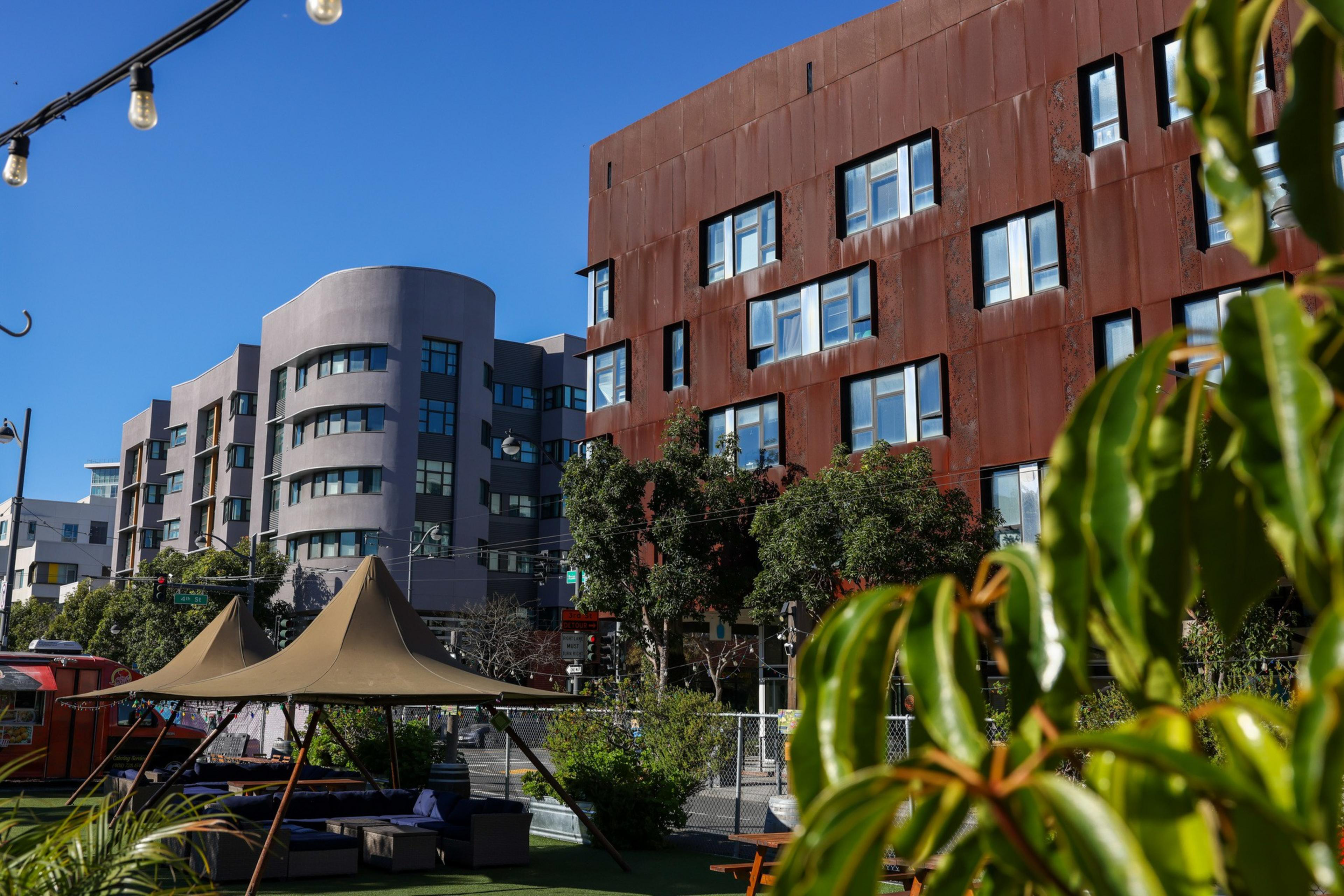 A modern urban scene features two distinctive buildings, one grey and curved, the other rust-colored. Trees, plants, and string lights add a touch of greenery.