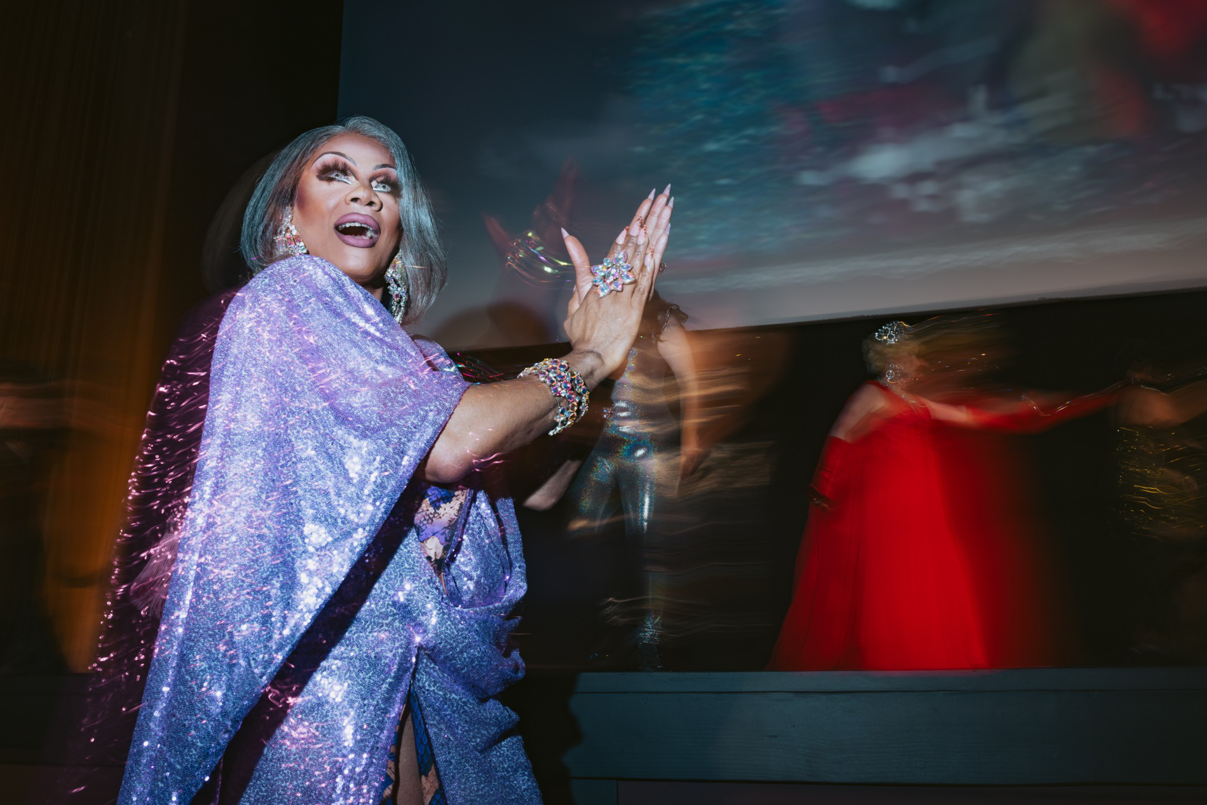 A person in a sparkling lavender outfit and dramatic makeup claps joyfully, with blurred figures and a dark backdrop behind them.